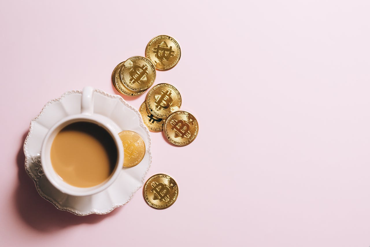 A cup of coffee with Bitcoin gold coins spread across a pastel pink surface.